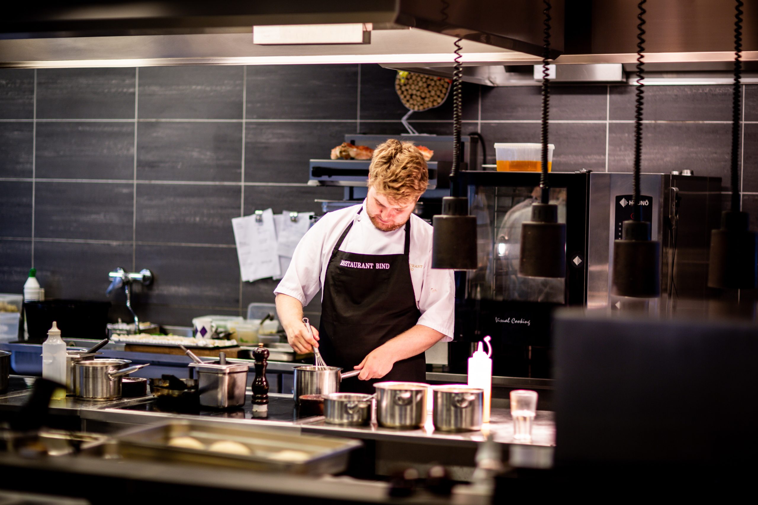 Young male chef preparing food in large commercial kitchen under fire suppression system.