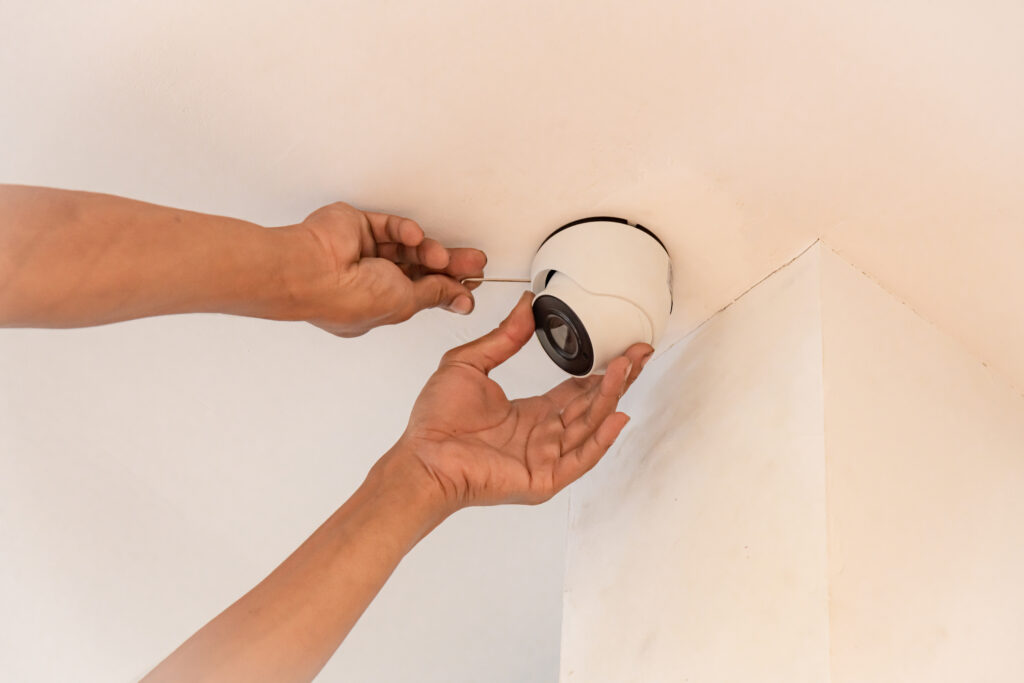 A pair of hands installing a small white round security camera on a white ceiling, near a white wall.