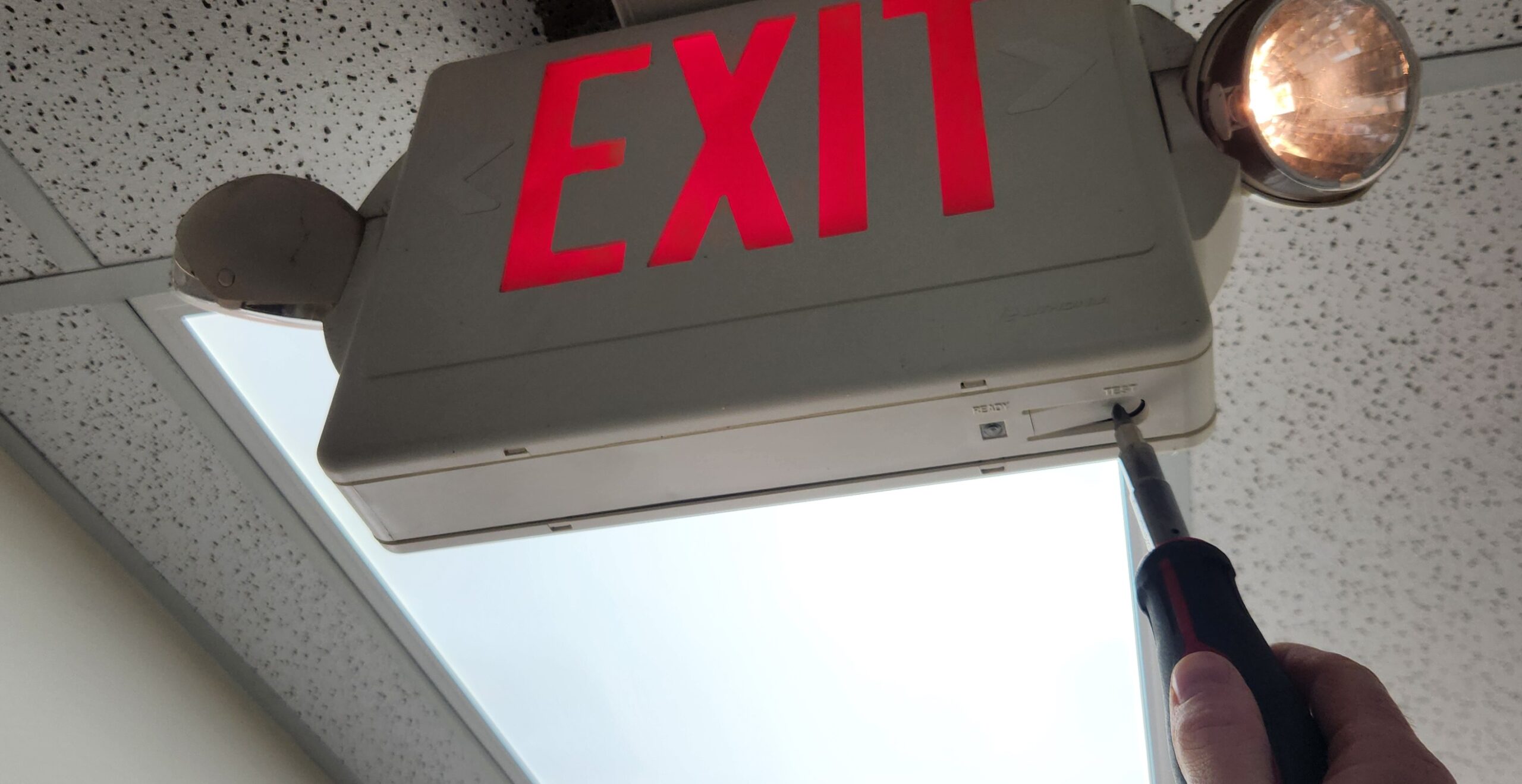 Close view of an exit sign on the ceiling of a commercial building; a technician's hand holding a tool to the side of the frame.